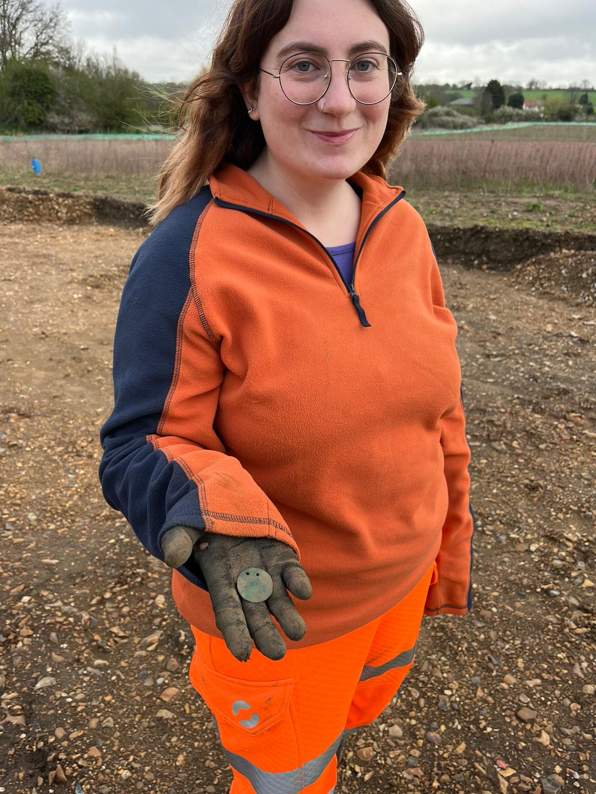 Archaeologist in an orange fleece holding a round metal disc pierced by two holes.