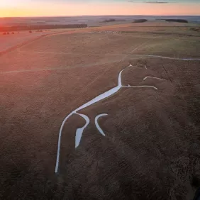 Aerial photo of the Uffington White Horse at sunrise, a prehistoric figure carved in the chalk of the downs. It is an abstract horse, with a long thin body, long tail. Its legs make it look as if it galloping over the hill. Image property of Hedley Thorne.