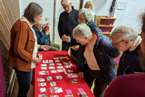 A female archaeologist stands behind a table with artefacts whilst various people examine them