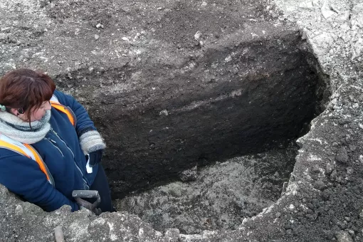 A female archaeologist stands in a large prehistoric storage pit cut into the natural chalk geology. The pit has been half excavated to show a half section of the soil fill.