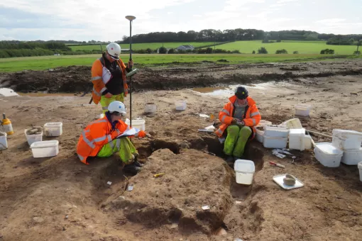 A group of archaeologists in high visibility uniform sit around a cremation burial carrying out recording of the feature