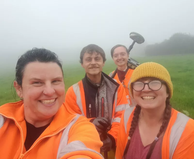 Image portraying a team of archaeologists completely soaked, on a very misty and wet site but all smiling together.